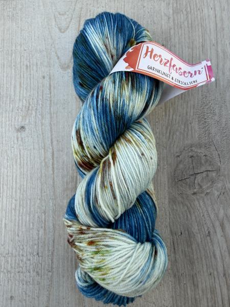 Handgefärbter Garnstrang in kühlen Blau- und Petroltönen mit cremefarbenen Abschnitten und feinen Sprenkeln in warmen Farben auf hellem Holzhintergrund.